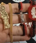 El significado espiritual de llevar un dije de la Santa Muerte: Una mano sostiene un dije dorado y dos pulseras con imágenes de la Santa Muerte, una roja y una negra, mostrando detalles de los accesorios.
