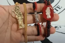 El significado espiritual de llevar un dije de la Santa Muerte: Una mano sostiene un dije dorado y dos pulseras con imágenes de la Santa Muerte, una roja y una negra, mostrando detalles de los accesorios.