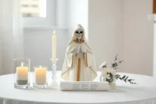 Niña Blanca: Una figura de la Santa Muerte vestida de blanco y dorado, acompañada de tres velas blancas encendidas, un arreglo floral blanco y un pergamino extendido con inscripciones, sobre una mesa blanca en un entorno iluminado.