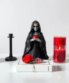 Oración Poderosa a la Santísima Muerte Negra para el Amor: Una estatua de la Santa Muerte Negra sosteniendo un corazón rojo, rodeada de cuatro velas encendidas, dos rojas y dos negras, con una rosa roja decorativa sobre una mesa blanca.